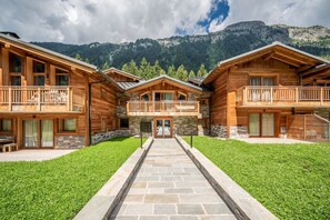 A wooden lodge with a central entrance, stone pathway, and balconies overlooking a mountainous landscape.