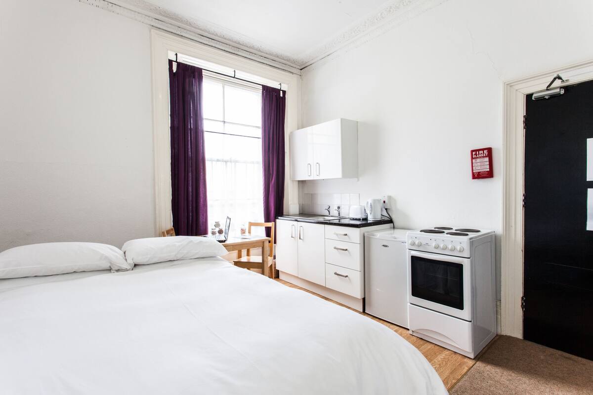A hotel room with a bed, a small kitchenette with a stove and coffee maker, a chair, and a window with purple curtains.