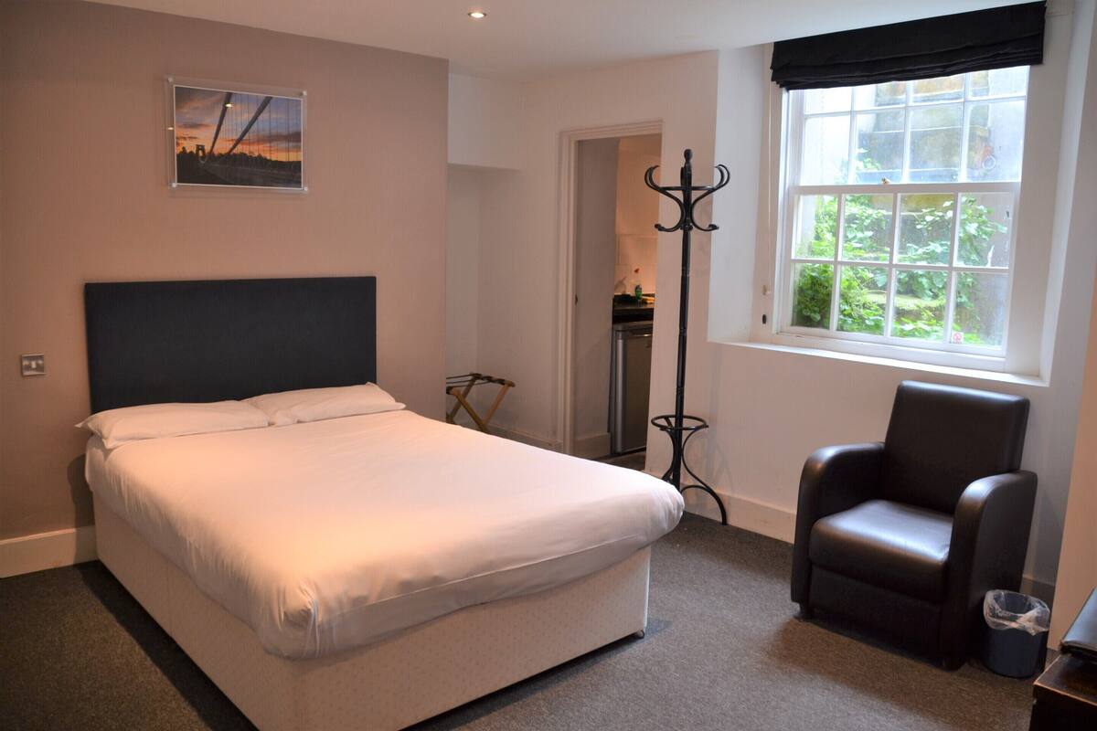 A bedroom with a bed, a chair, a coat rack, and a window with a view of greenery.