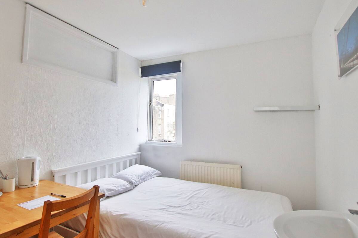 A small, white room with a single bed, a wooden chair, a table, and a window.