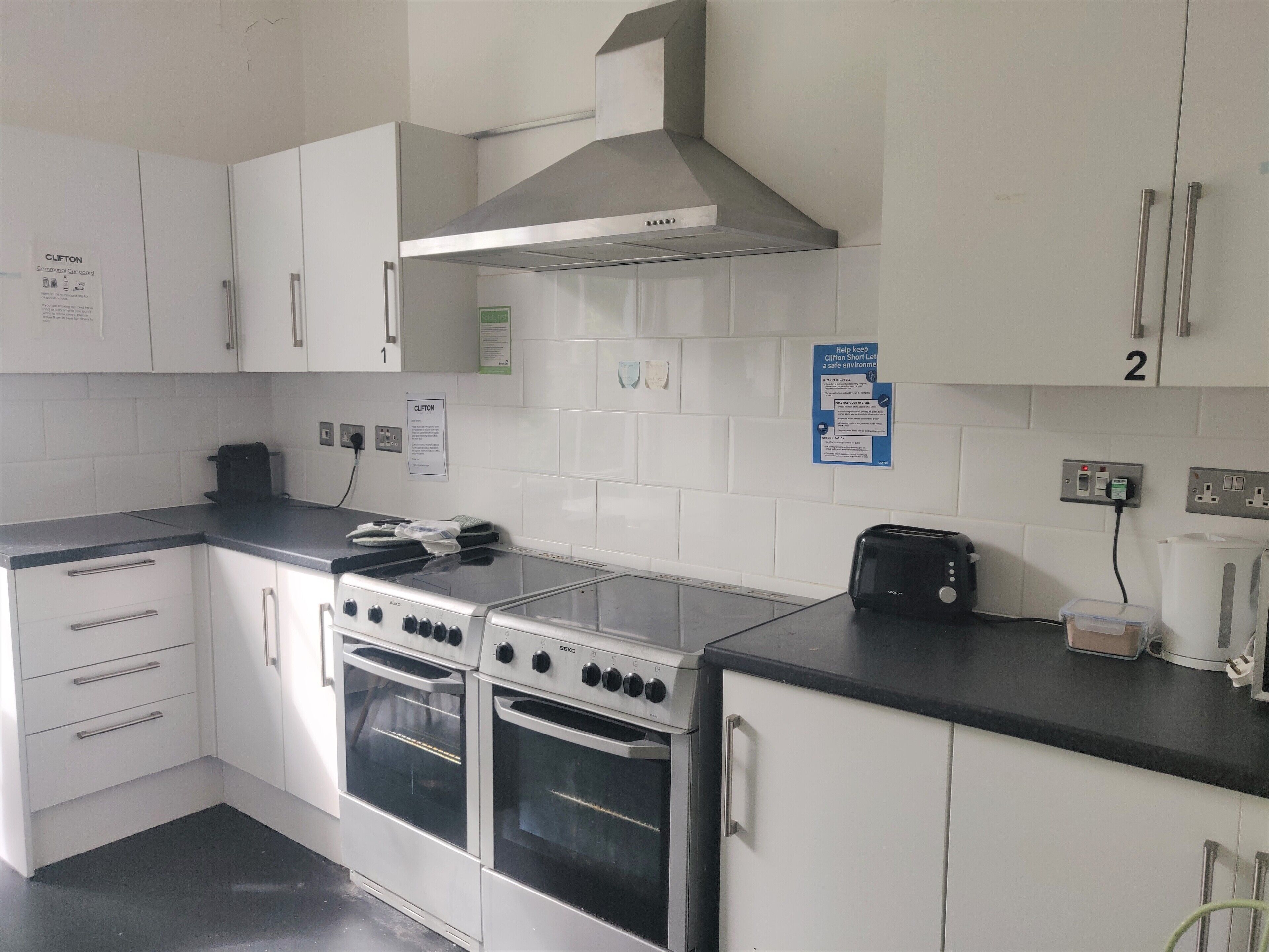A kitchen with white cabinets, a stainless steel range hood, a toaster, and a kettle. There are two ovens and a countertop with a kettle and a toaster.