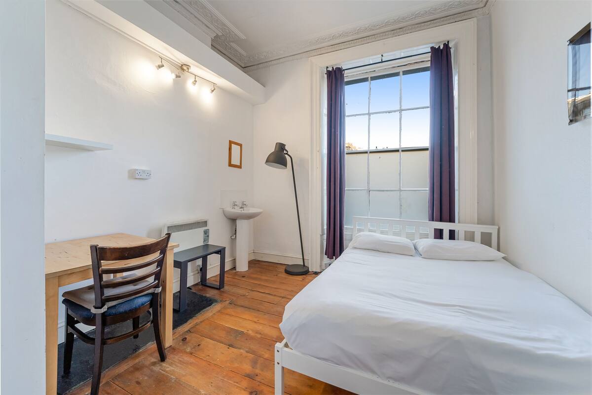 A bedroom with a bed, a desk with a chair, a bedside table, a lamp, a window with curtains, and a view of the sky.
