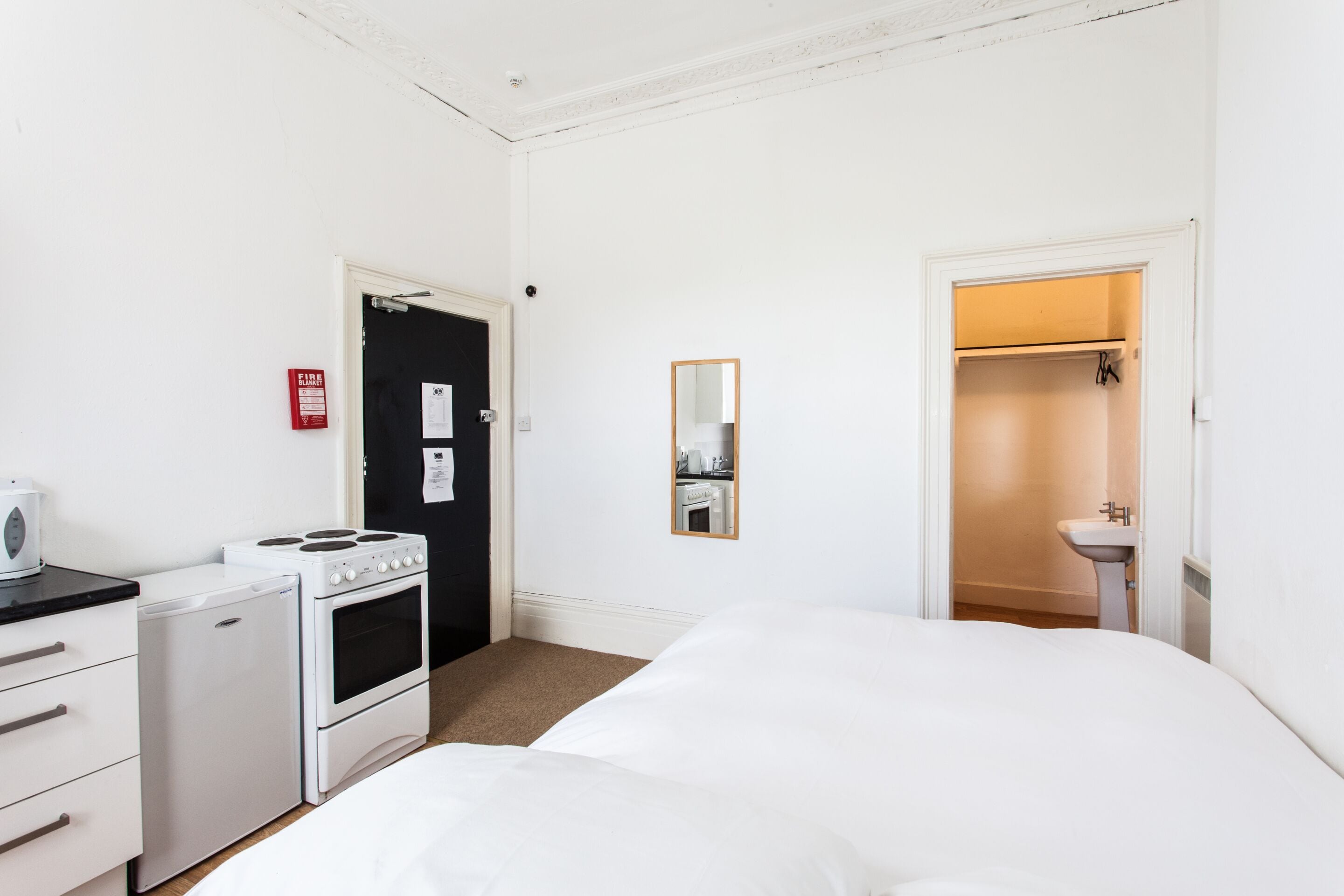 A hotel room with a bed, a small kitchenette with a stove and refrigerator, a mirror, and a bathroom with a sink and towel rack.