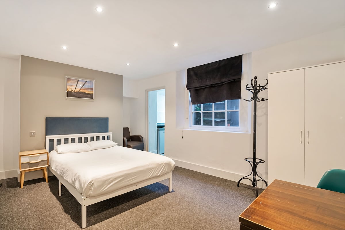 A bedroom with a bed, a nightstand, a chair, a wooden table, a coat rack, and a wardrobe.