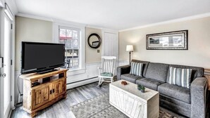 A living room with a flat-screen TV on a wooden cabinet, a grey sofa with striped pillows, a white coffee table, and a white chair. - The Breakwaters Hyannis - Your Home Away From Home! RPAM046 (Hyannis)