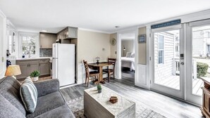 A modern living room with a gray sofa, a coffee table, and a dining area with a wooden table and chairs. A kitchen with white cabinets and a refrigerator is visible in the background. - The Breakwaters Hyannis - Your Home Away From Home! RPAM046 (Hyannis)