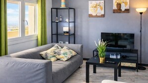 A modern living room with a grey sofa, a black TV stand, a small table, and a bookshelf. - Ocean View Apartment • Sunny Balcony • Near the Coast (Antigua)