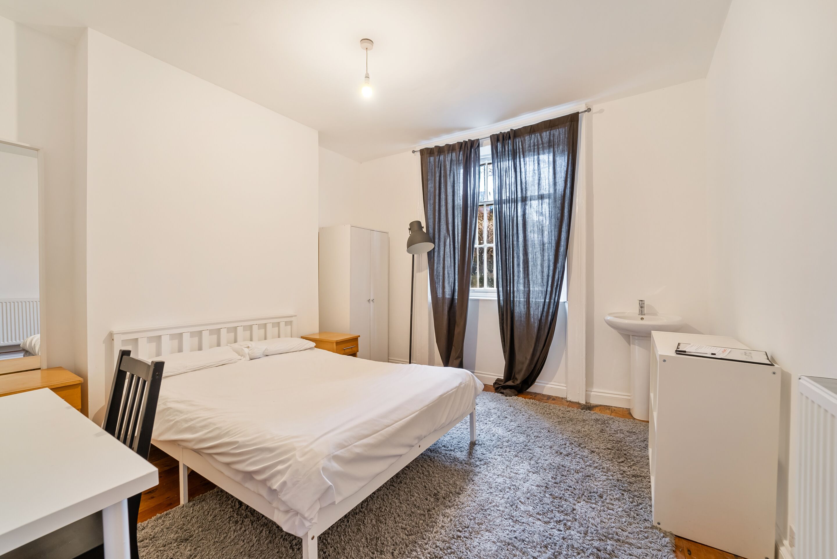 A bedroom with a single bed, a desk, a chair, a radiator, a wardrobe, a floor lamp, a sink, and a refrigerator.