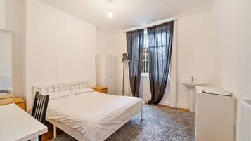 A bedroom with a single bed, a desk, a chair, a radiator, a wardrobe, a floor lamp, a sink, and a refrigerator.