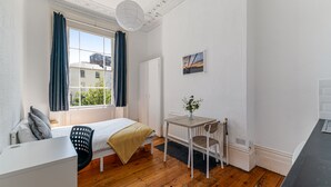 A small, tidy room with a bed, a desk, and a chair. There is a window with curtains, a radiator, and a small table with a plant.