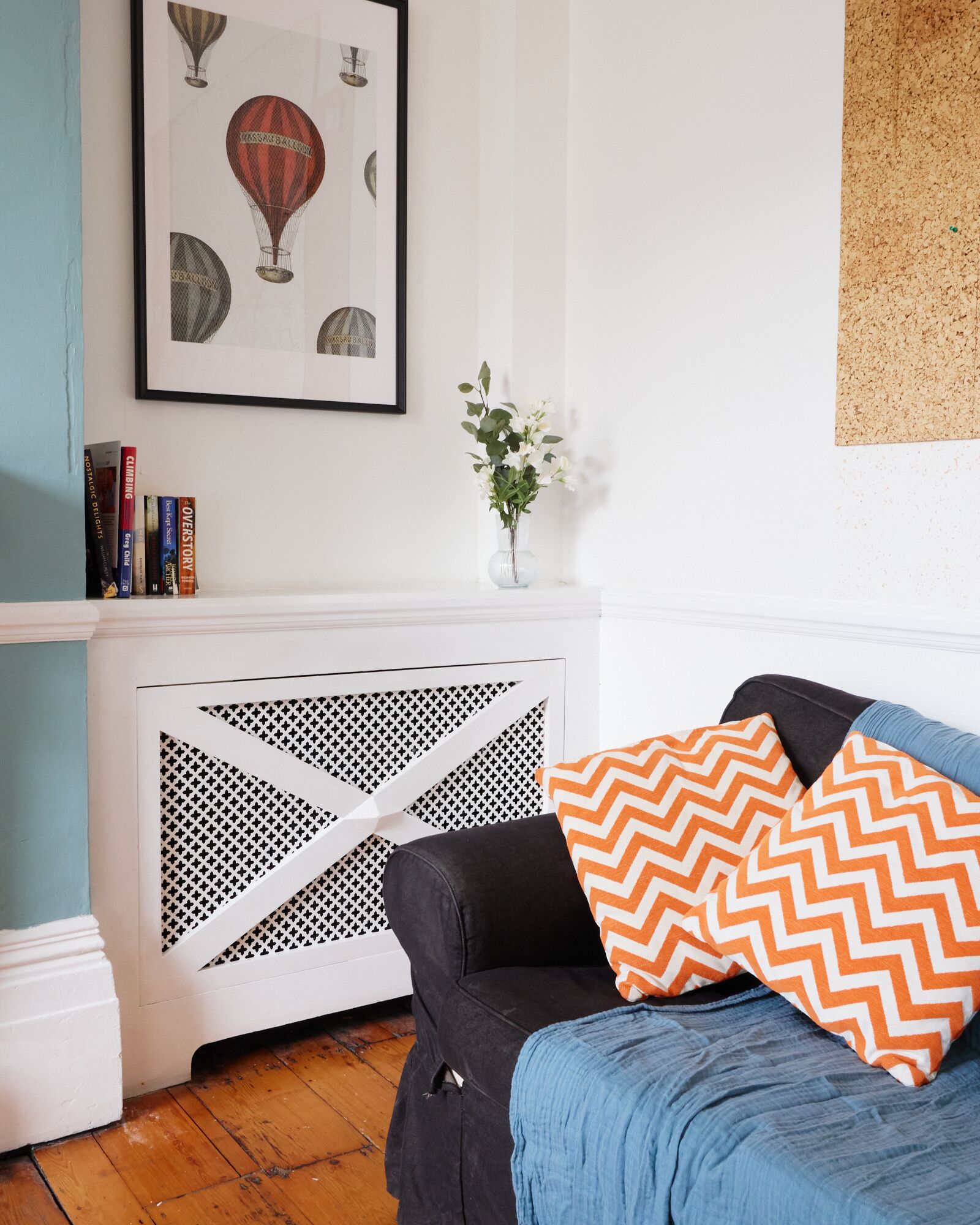 A room with a black sofa, a blue blanket, a chevron-patterned pillow, a white radiator with a geometric design, a framed picture of hot air balloons, and a vase with white flowers.