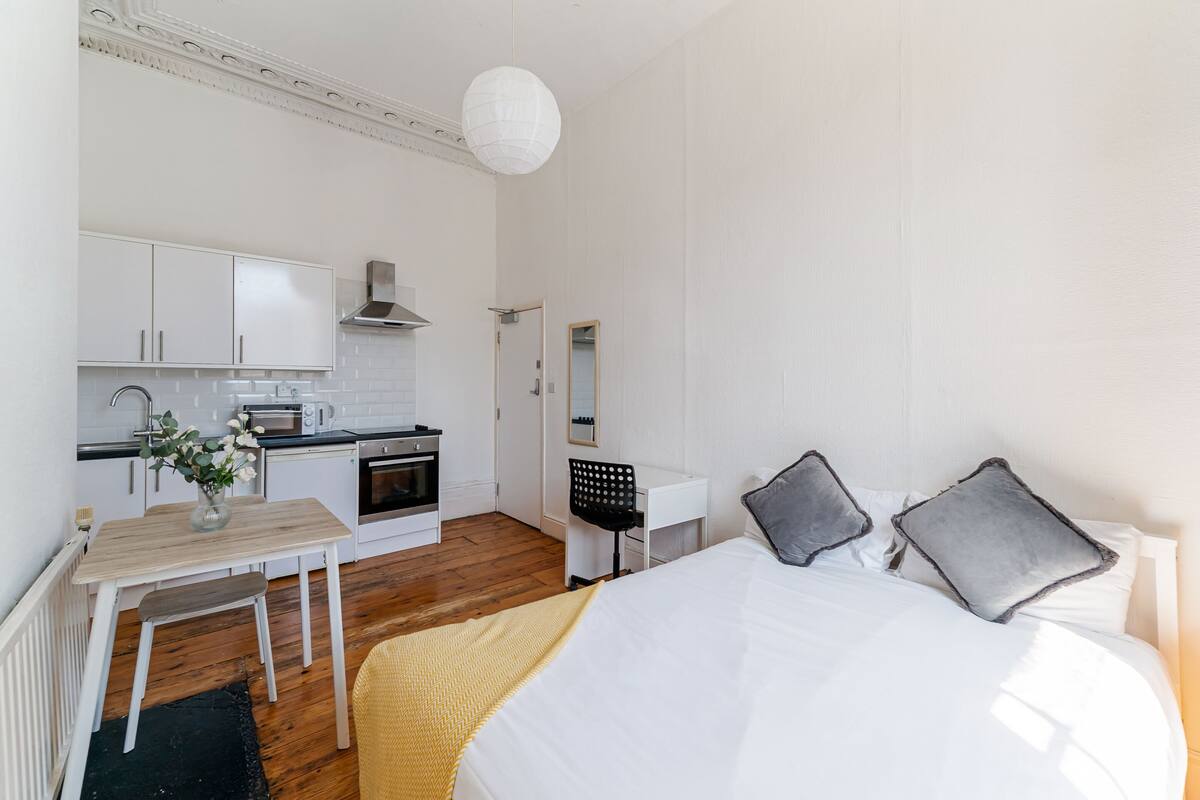 Een compacte, goed verlichte kamer met een bed, nachtkastje en een kleine kitchenette.