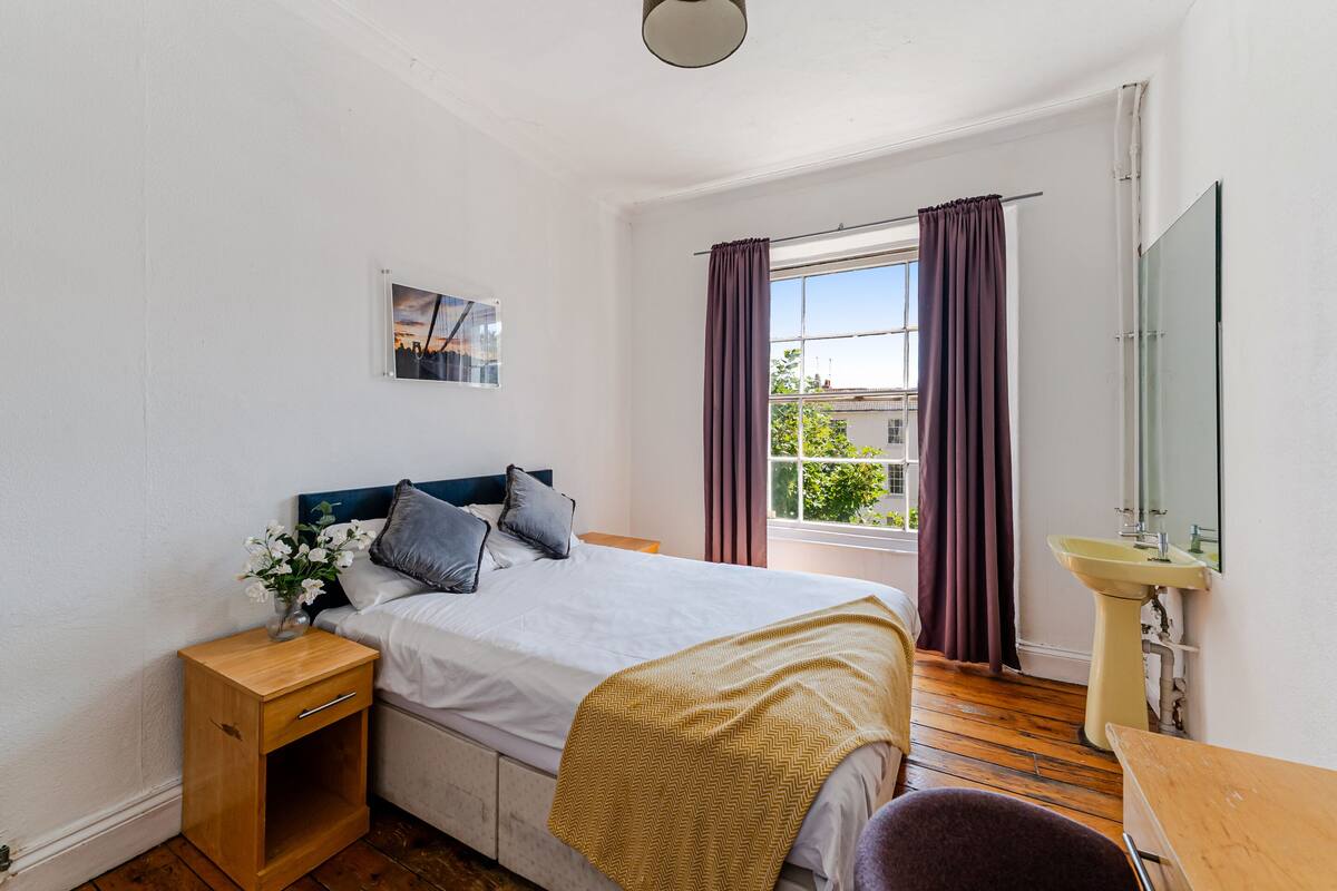 Een slaapkamer met een bed, nachtkastje, raam met gordijnen en een spiegel.