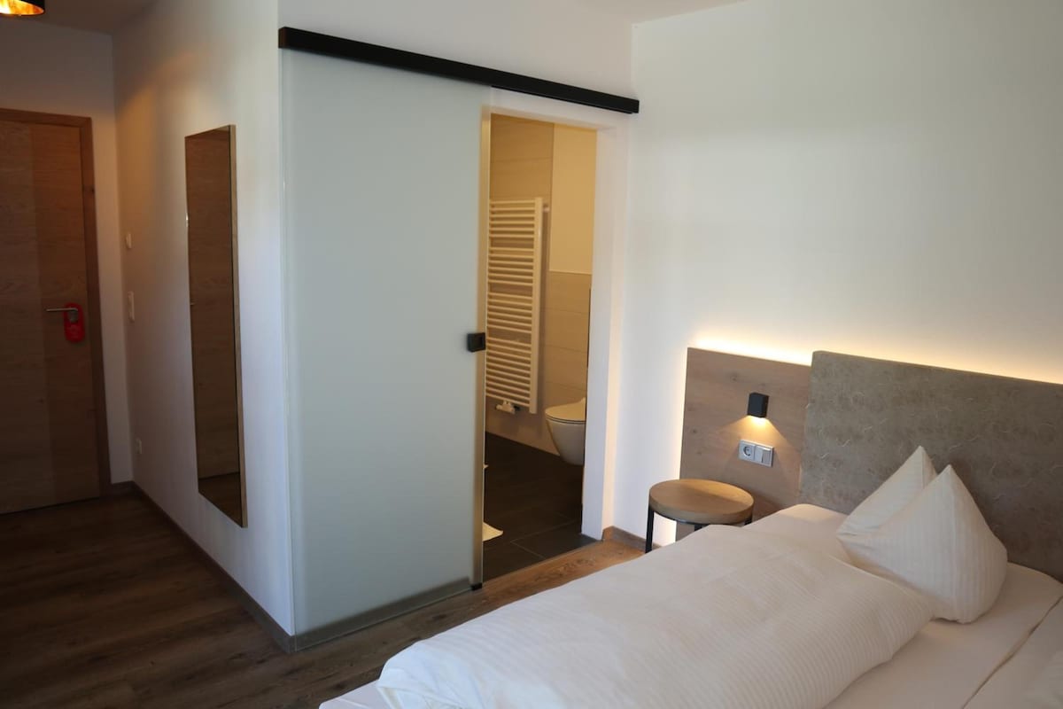 A hotel room with a bed, a nightstand, a wall-mounted light, and a door leading to a bathroom.