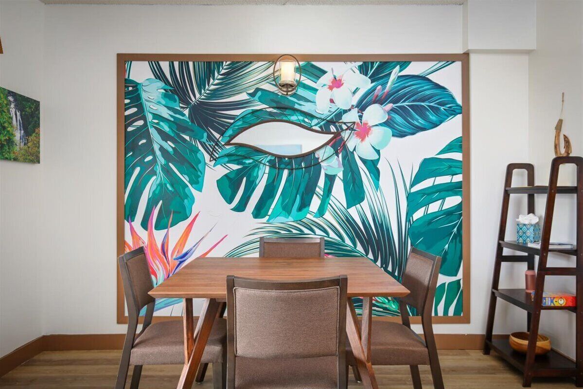 A dining area with a wooden table and chairs, a large tropical leaf and flower mural on the wall, and a small shelf with decorative items.