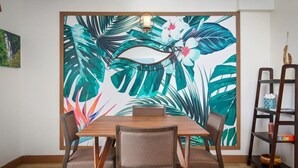 A dining area with a wooden table and chairs, a large tropical leaf and flower mural on the wall, and a small shelf with decorative items.