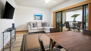 A modern living room with a sectional sofa, a flat-screen TV, a dining table, and a balcony with outdoor furniture.