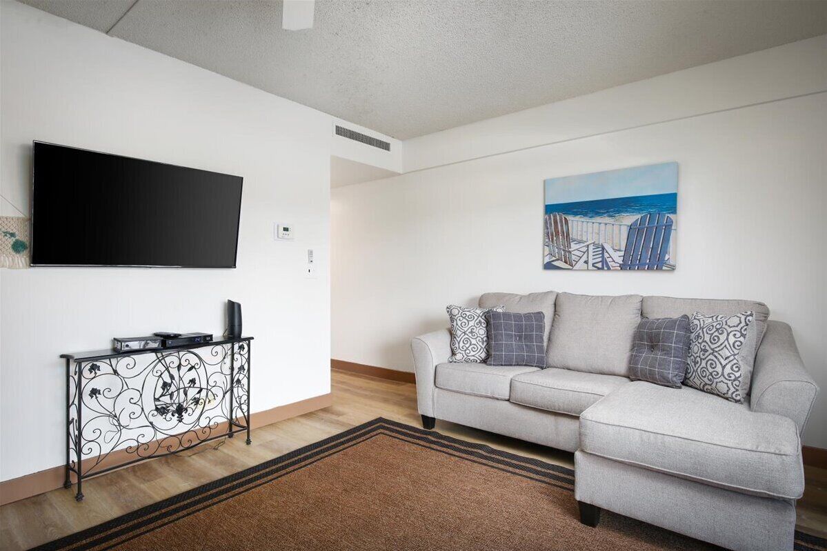 A living room with a flat-screen TV, a sofa with patterned cushions, a decorative metal console table, and a painting of beach chairs.