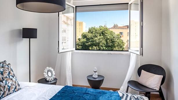 A bedroom with a bed, a chair, a small table, a lamp, and a window with a view of buildings and trees. - Sapphire Apartment, AC, Wifi !new! (Lisboa)