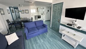Ein modernes Wohnzimmer mit einem blauen Sofa, einem Essbereich mit Tisch und Stühlen, einer Küche mit weißen Schränken und einem Fernseher auf einem weißen Schrank.