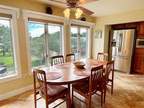 A dining area with a wooden table and chairs, large windows with a view of greenery, and a kitchen with stainless steel appliances.