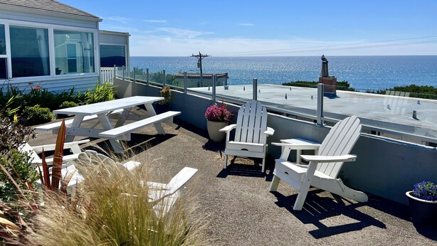 A patio with white Adirondack chairs and a picnic table overlooking a scenic ocean view.