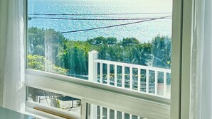 A room with a large window overlooking a coastal view, a white railing, and a view of the sea and greenery.