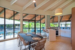 A spacious dining area with a large table and chairs, a kitchen with white cabinets, and large windows offering a view of greenery.