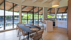 A spacious dining area with a large table and chairs, a kitchen with white cabinets, and large windows offering a view of greenery. - Stylish and Spacious Condo - A/C in 2 Bedrooms, Huge Lanai, Great Location (Princeville)