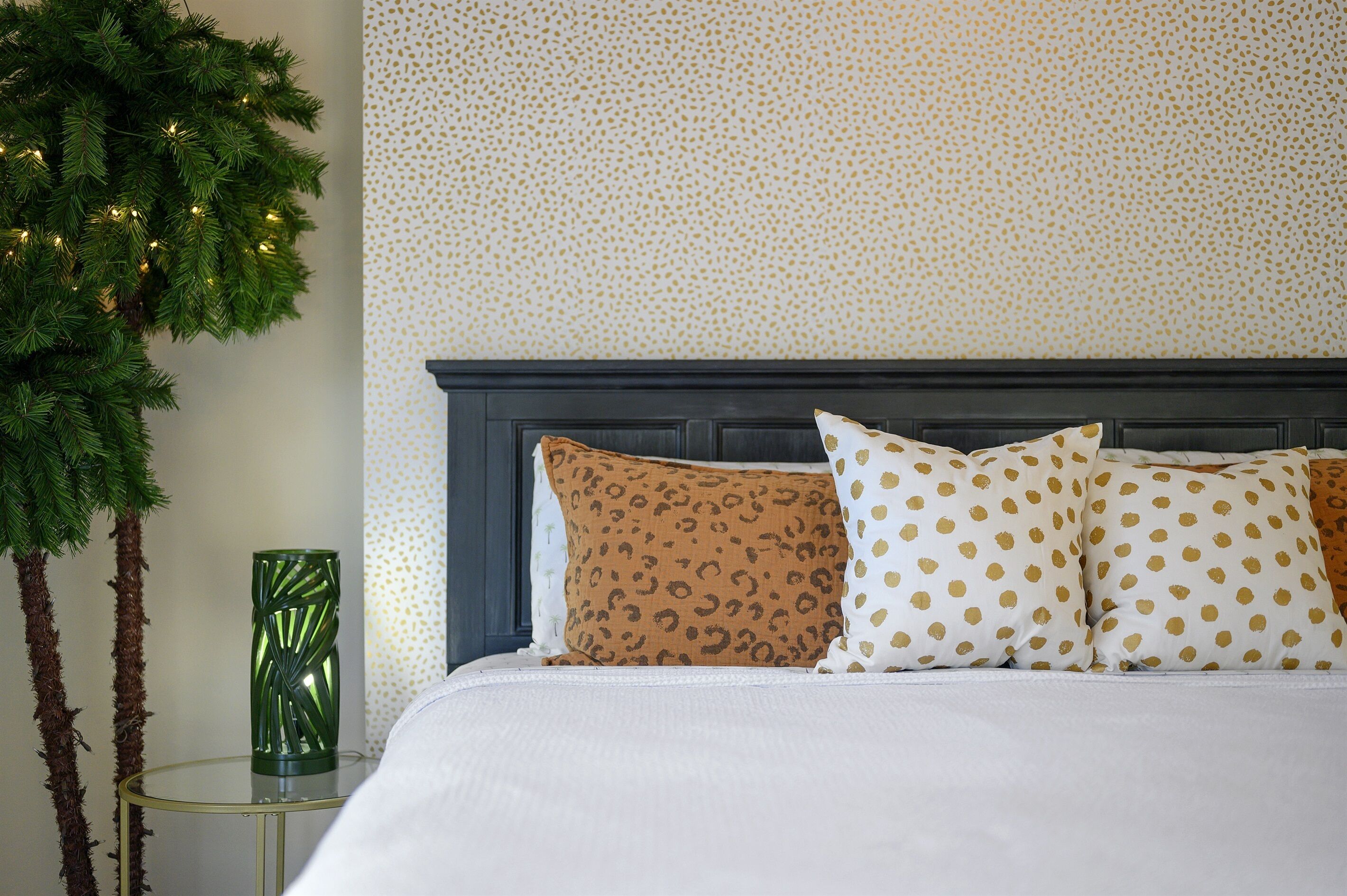 Een net opgemaakt bed met patroonkussens, een klein nachtkastje met een groene vaas, en een muur met een gespetterd patroon.