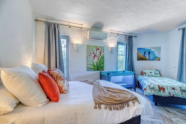 A bedroom with a large bed, a smaller bed, a desk, and a chair. There are two windows with curtains, an air conditioning unit, and artwork on the walls.