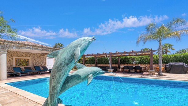 En pool med en delfinskulptur, solstolar och en pergola med parasoller.