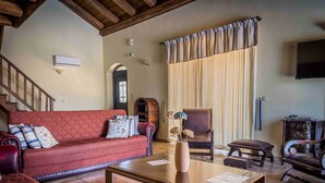 Una sala de estar acogedora con un sofá rojo, una mesa de centro de madera y una silla. Se observa una escalera con barandillas de madera, una chimenea y un televisor montado en la pared.