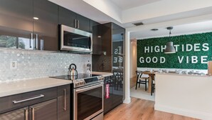 Una cucina moderna con mobili scuri, un forno a microonde e un forno in acciaio inossidabile. C'è un'area pranzo con tavolo e sedie, e una parete con la scritta 'HIGH TIDES GOOD VIBE'.
