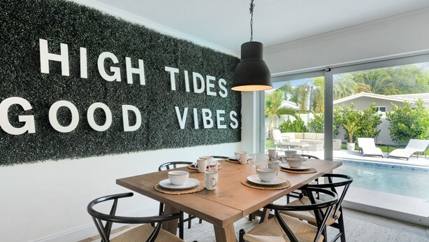 Zona pranzo con vista piscina, parete nera con scritta 'HIGH TIDES GOOD VIBES' e tavolo in legno con sedie.