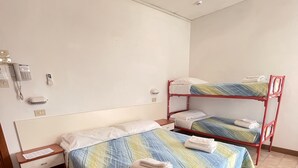 A single bed with a colorful bedspread, a wooden nightstand, a red bunk bed, and a wall-mounted phone.
