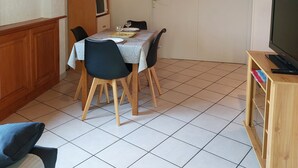 A dining area with a table set for two, a television on a wooden stand, and a kitchen area in the background. - Appartement (Gueberschwihr)
