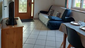 A cozy living room with a wooden ceiling, a sofa with cushions, a TV on a wooden stand, a dining table with chairs, and a window with curtains. - Appartement (Gueberschwihr)