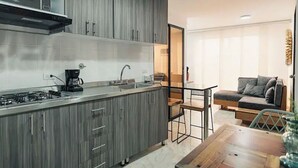 A modern kitchen with grey cabinets, a built-in microwave, and a gas stove. There is a dining area with a wooden table and chairs, and a grey sofa in the living room. - Hermoso y Cómodo Apartamento en El Poblado!!! (Medellín)