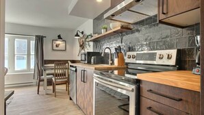 A modern kitchen with a built-in oven, a microwave, and a stove. There is a wooden dining table with chairs, and a window with curtains. - Unique & Sunny Top Floor Old Quebec Gem (Québec City)