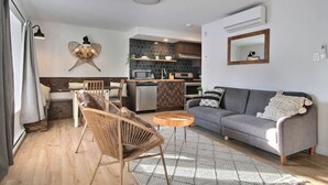 A modern living room with a grey sofa, a wooden coffee table, and a wicker chair. A kitchen is visible in the background with dark tiles and wooden cabinets. - Unique & Sunny Top Floor Old Quebec Gem (Québec City)