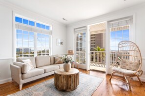 A spacious living room with a beige sofa, a round wooden coffee table, and a wicker chair. - Pier Point | Ocean View | Walk To Beach (Folly Beach)