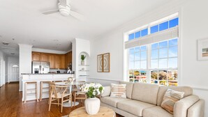 Amplia sala de estar con un sofá color beige, una mesa de comedor de madera y sillas. Un jarrón blanco con flores está sobre un taburete de madera. Un ventanal ofrece vistas de la ciudad.