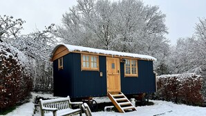 Una cabaña pequeña y azul oscuro con puerta y ventanas de madera, rodeada de árboles cubiertos de nieve y un banco al frente.