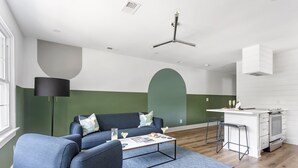 Een moderne woonkamer met een blauwe bank, een salontafel en een keukenhoek met een wit aanrecht en barstoelen.