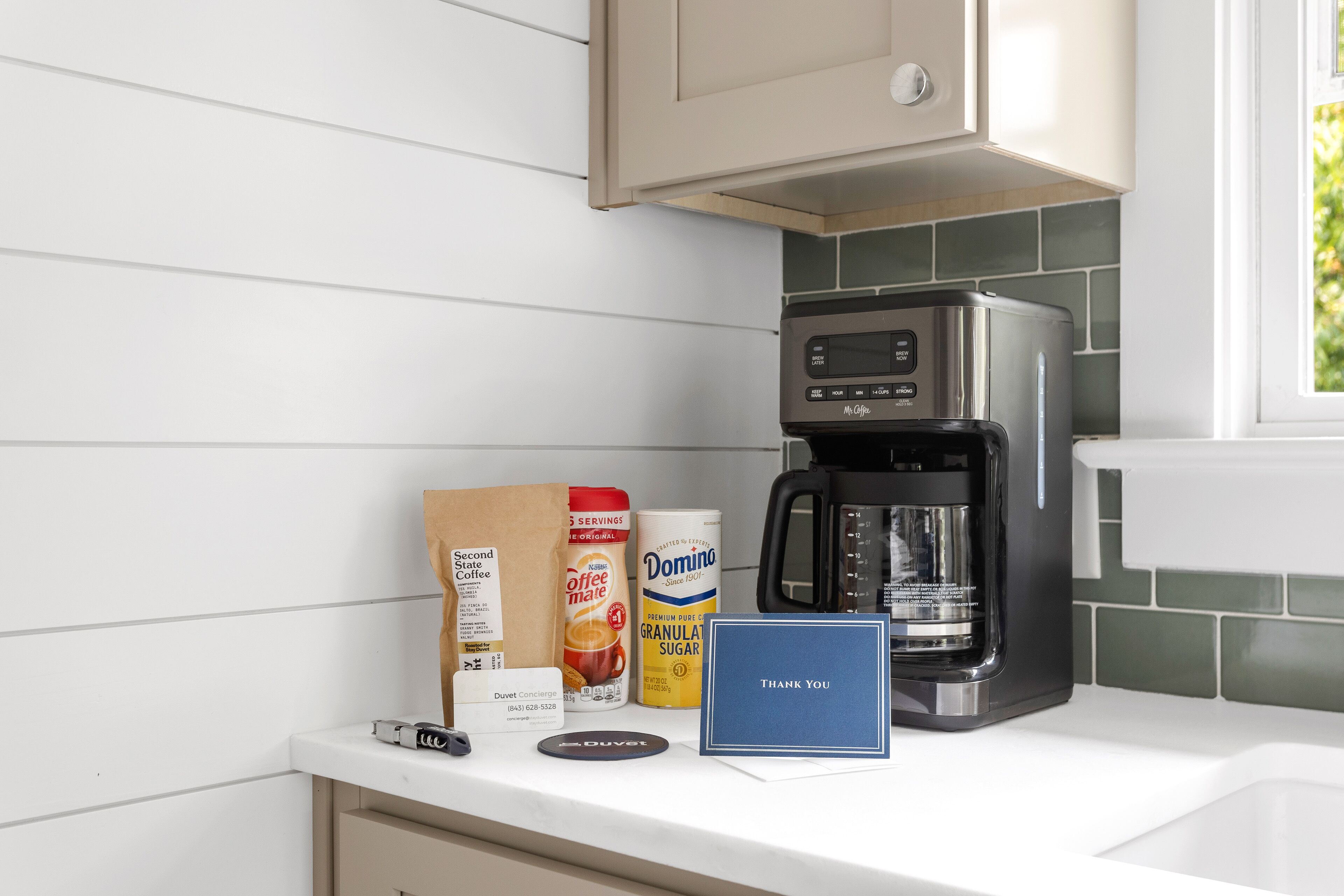 Una encimera de cocina con una cafetera, un paquete de café Domini, un bote de café Domini, una tarjeta Duvet Concierge y una tarjeta de agradecimiento.