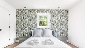 Een slaapkamer met behang met een geometrisch patroon, een bed met witte lakens, twee nachtkastjes en een raam met uitzicht op bomen.