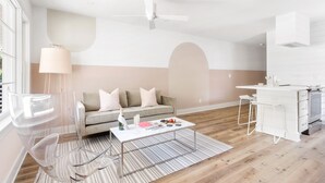 Una sala de estar moderna con un sofá, una mesa de centro, una silla transparente y una zona de cocina con encimera blanca y taburetes de bar.