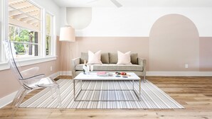 Una sala moderna con una silla de vidrio transparente, un sofá con cojines, una mesa de centro y un amplio ventanal con vistas a zonas verdes.