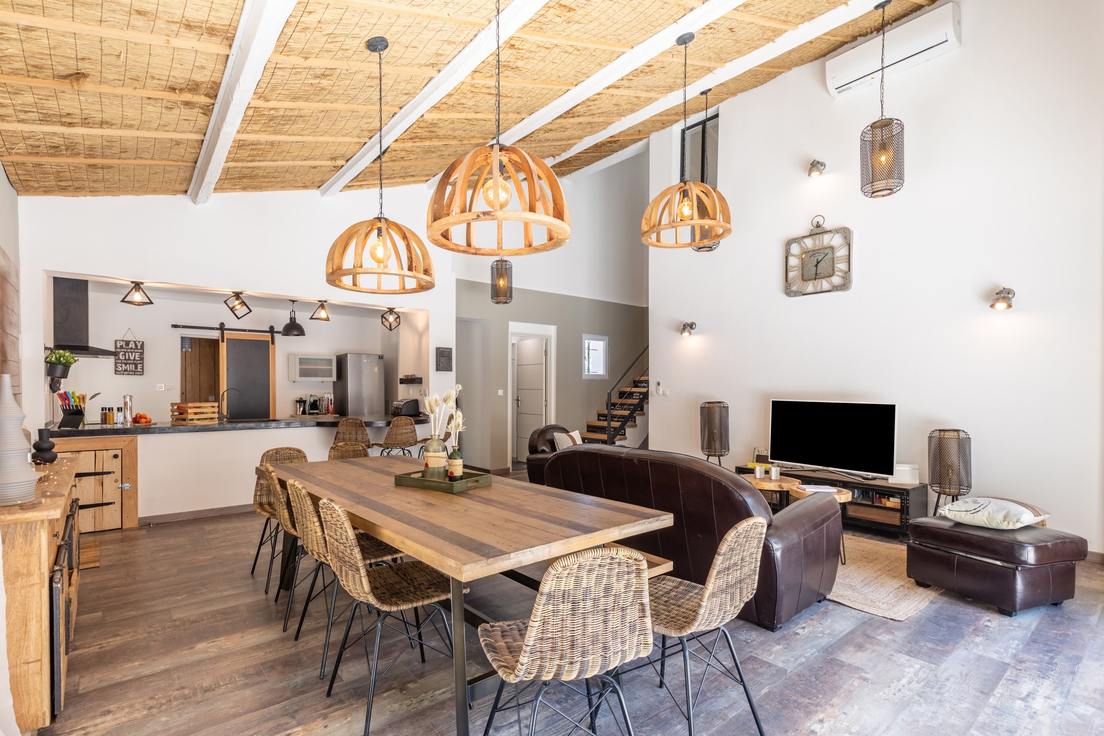 A spacious living area with a wooden dining table, wicker chairs, a leather sofa, and a TV.